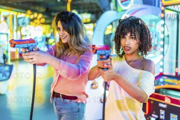 Two women laughing and enjoying their free time while playing colorful arcade shooting games, immersed in fun and friendly competition