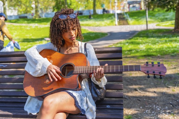 Musician concentrating while playing acoustic guitar sitting on a bench in a green city park during a sunny spring day