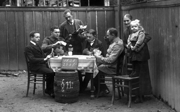 Round of cards, men playing cards, 1910, Group of people sitting outside at a table playing cards. A child is held in the arms of a woman, historical photo