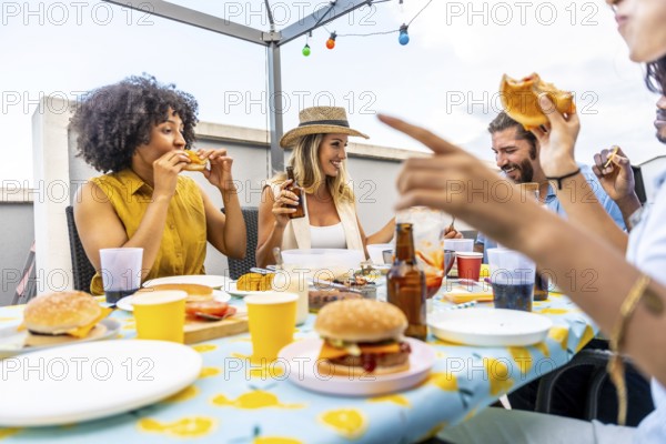 Happy friends enjoying burgers and beer, celebrating togetherness on a rooftop with string lights