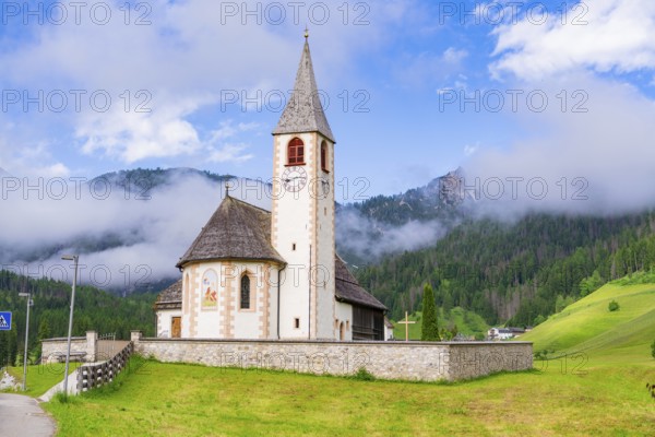 Church with tower and clock, surrounded by meadows and mountains in an alpine landscape, Dolomites, South Tyrol, Italy