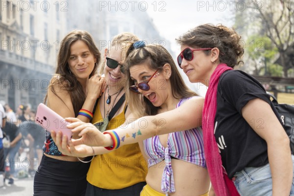 Four young women wearing rainbow bracelets are taking a selfie with a smartphone during a gay pride parade, making funny faces and showing their enthusiasm