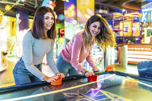 Two female friends enjoying their free time playing air hockey in a colorful and modern amusement arcade