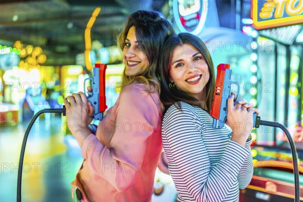 Two joyful women standing back to back, holding toy guns and enjoying playful moments in a vibrant, brightly lit amusement arcade