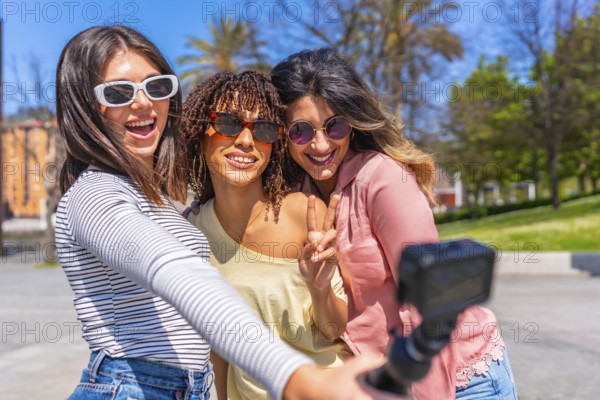 Three cheerful young women making peace sign and taking selfie with action camera during summer vacation in the city