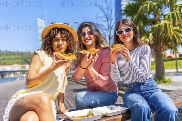 Three women enjoying pizza slices while sitting on a bench in a sunny city park, sharing laughter and savoring a delightful summer day