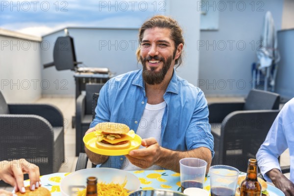 Happy man holding a burger during a rooftop barbecue party with friends, enjoying good food and company
