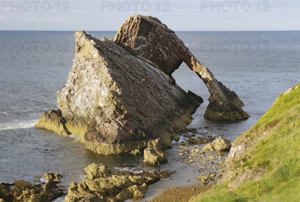 Large rock formation in a calm sea illuminated by the setting sun, Remarkable rock formation jutting into the sea under a blue sky with few clouds, Bow Fiddle Rock, Aberdeenshire, Buckie, Scotland, United Kingdom
