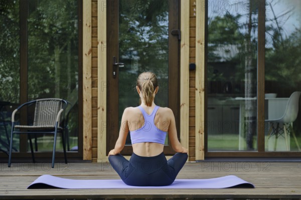 Back view of a woman sits in lotus position on a yoga mat placed on a wooden terrace of tiny house