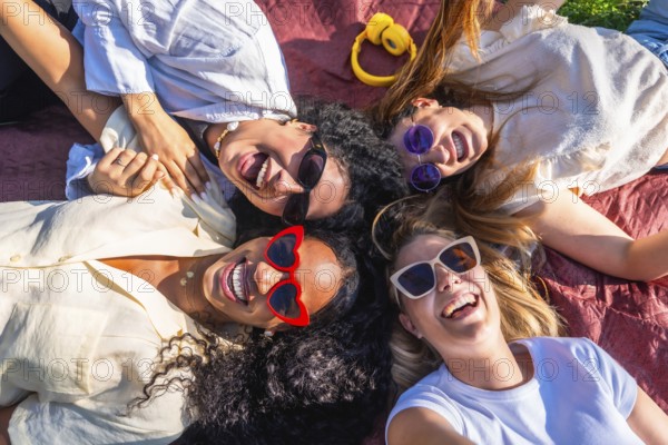 Four cheerful young women wearing sunglasses are lying together on a blanket in a park, happily laughing and enjoying their time together