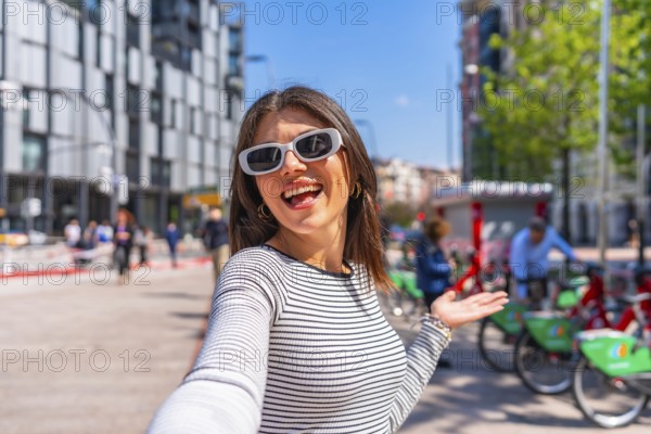 Happy young woman taking a selfie in the city during summer, gesturing to show the urban landscape