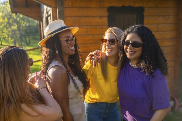 Four multi ethnic female friends enjoying a sunny day, wearing sunglasses and laughing together near a wooden house in the countryside