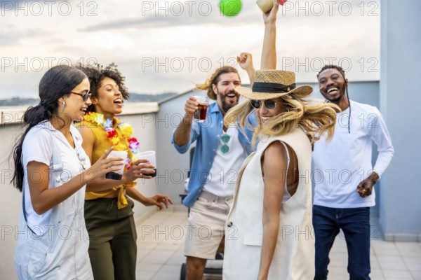 Cheerful multi ethnic friends enjoying cocktails while dancing joyfully at a vibrant rooftop party, celebrating summer under the sunset