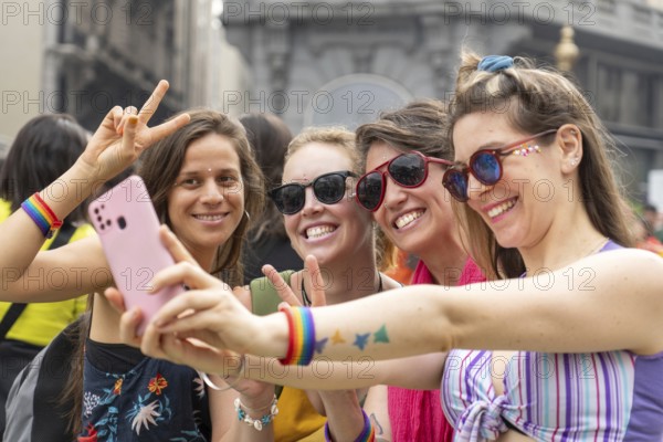 Four joyful women wearing colorful rainbow bracelets are capturing a selfie with a smartphone during a vibrant pride parade, flashing peace signs and beaming with happiness