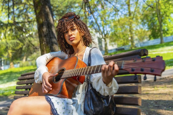 Young woman sitting on a park bench, playing an acoustic guitar while enjoying a sunny day filled with music and relaxation outdoors