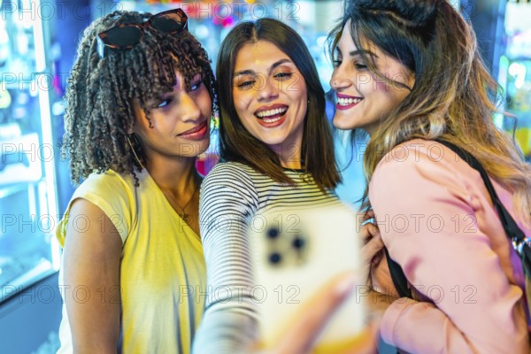 Three cheerful young women enjoying a fun moment together, taking a selfie with a smartphone in a vibrant amusement arcade at night