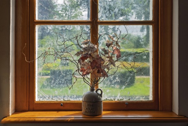 A ceramic vase with dried branches stands on a sunny windowsill in front of an old window with a garden view