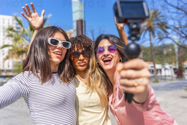 Three cheerful women holding action camera and waving at camera while vlogging their summer vacation in a sunny city