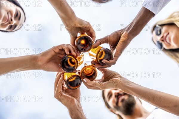 Group of diverse friends are holding and toasting glass bottles of beer together, celebrating under a cloudy blue sky