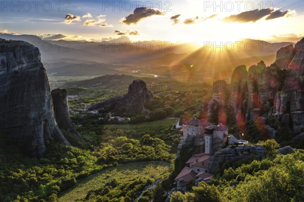 Iconic view of Meteora valley at sunset. Sun setting behind distant mountains, sun rays fall on valley, cliffs, rocks, clifftop Moni Agias Varvaras Roussanou nunnery, blue sky and beautiful clouds. Natural lens flares. Greece, UNESCO World Heritage