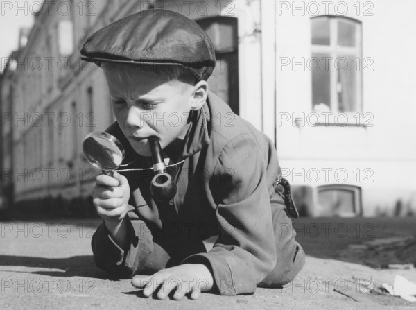 Playing detective. A young boy dressed as a typical looking detective with a magnifying glass and a pipe, is playing outside. Sweden 1961