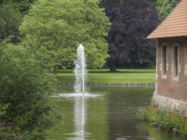 Fountain in a green park next to a brick building, with large trees and calm water, havixbeck, westphalia, germany