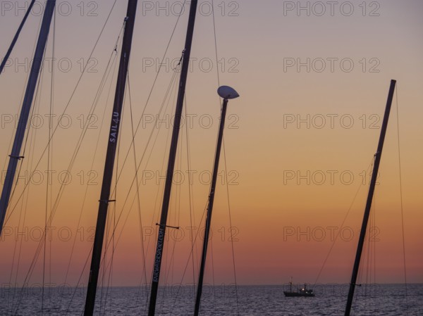Masts in the foreground and a small boat in the sea at sunset, the sky is bathed in orange tones, De Haan, Flanders, Belgium