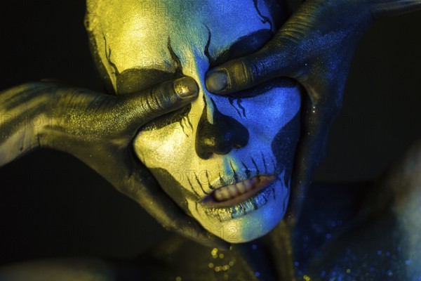 Isolated on black, closeup picture, toned yellow and blue, spooky young blonde caucasian pretty woman with scull body art, closed grey eyes, closes her eyes with her thumbs