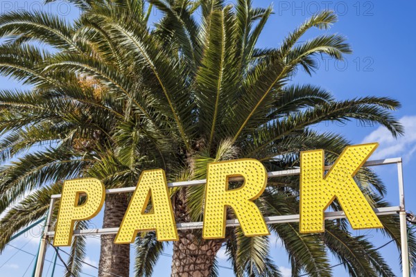 Entrance to an amusement park with big yellow lights sign and palm trees with bright blue sky on the background. Summer vacations concept