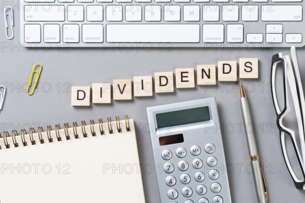 Dividends payment concept with letters on wooden cubes. Still life of office workplace with supplies. Flat lay grey surface with keyboard, notepad and calculator. Capital savings and investment
