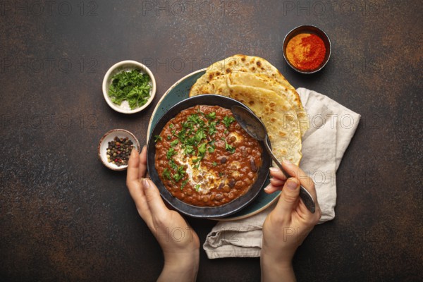 Female hands holding a bowl and eating traditional Indian Punjabi dish Dal makhani with lentils and beans served with naan flat bread, fresh cilantro on brown concrete rustic table top view, food photography, food photography