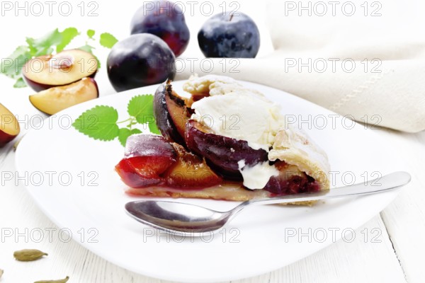 Piece of sweet pie with plum, sugar, cardamom and ice cream in a plate, sprigs of green mint, towel on light wooden board background