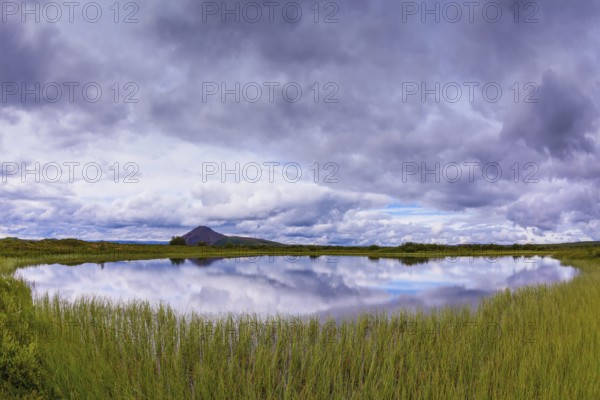 Small lake surrounded by green meadow. In the smooth water of cold lake reflects cloudy sky. Summer Iceland