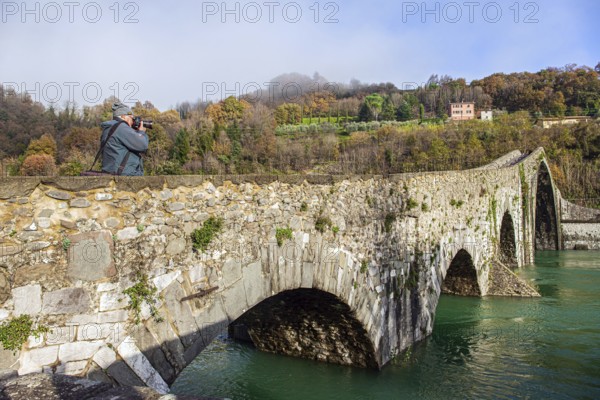 Elderly tourist photographs a magnificent landscape. Ponte della Maddalena Bridge. Cold green waters of the Sercchio River. Italy. Province of Tuscany. Cold windy winter day