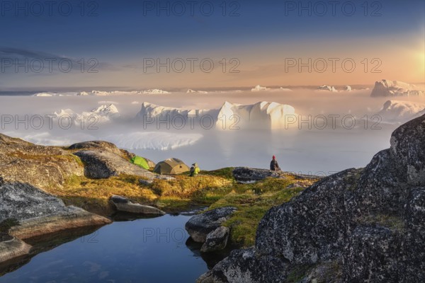 Large glaciers in the bay by the rocky shore in the light of the setting sun with fog over the water, woman in a red cap
