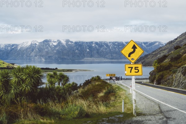 Road with danger signs leads along the lake in front of snow-covered mountains, Wanaka, New Zealand