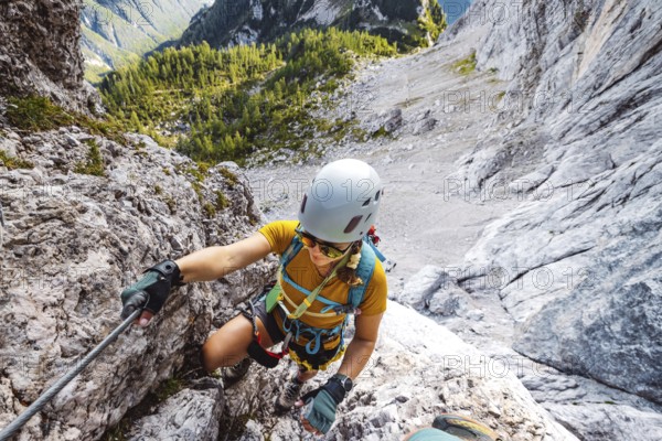 Caucasian woman mountaineer climbing up the mountain on via ferrata trail, high up in the mountain peaks. Woman wearing a protective head gear and uses a safety harness while she climbs