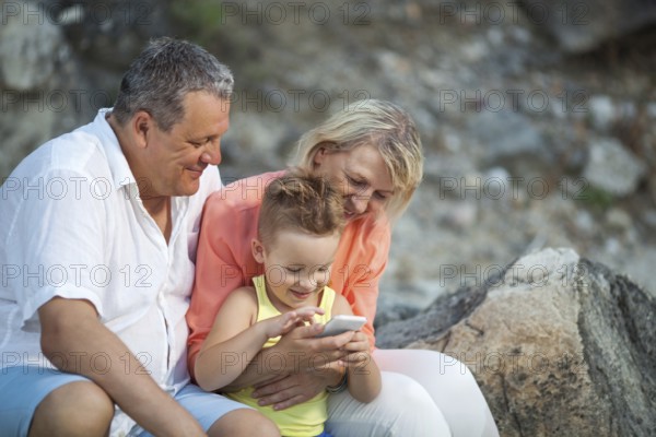 Happy grandparents and grandson with smartphone outdoor. Grandmother and grandfather watching the boy playing with mobile