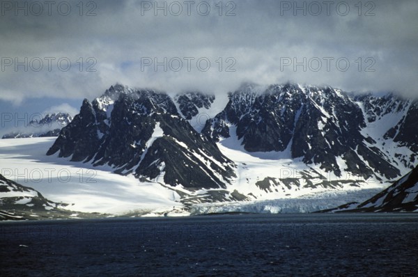 Snow-covered and glaciated rugged mountains surround the Magdalenefjord on Spitsbergen. Glaciers slide from the mountain flanks into the dark blue waters of the fjord. The mountain peaks are obscured by high cloud fields. Tiny in the arctic landscape, a cruise ship in front of the edge of the glacier. Snowcovered and glaciated rugged mountains surrounding the Magdalene Fjord on Spitsbergen. From the mountain flanks, glaciers slide into the dark blue water of the fjord. In the arctic landscape, a tiny cruise ship is off the drip line of the glacier
