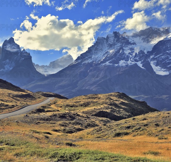 The gravel road is bent between yellowed hills and go to the snow-covered rocky mountains. Midday landscape in the national park Torres del Paine, Chile