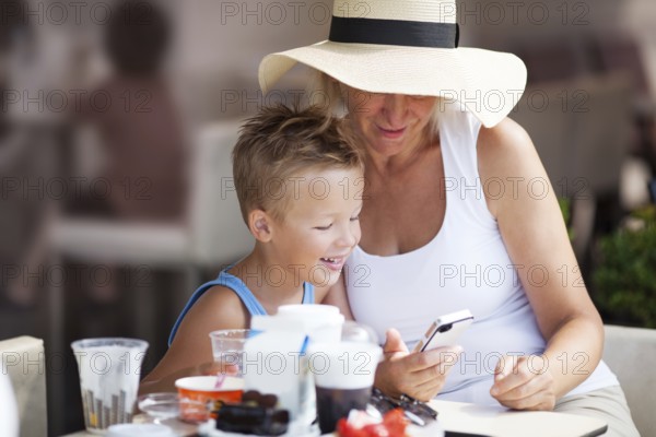 Smiling Mature Woman and Young Boy On Vacation Using Cell Phone Together on Outdoor Restaurant Patio