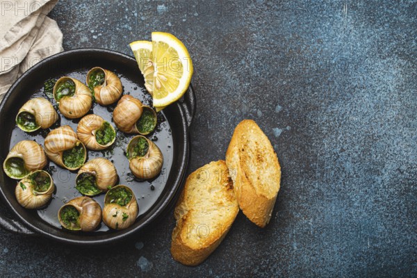 Escargots de Bourgogne Snails with Garlic Butter and Parsley in black cast iron pan with Lemon and Toasted Baguette Slices on rustic stone background top view, traditional French Delicacy, copy space, food photography, food photography