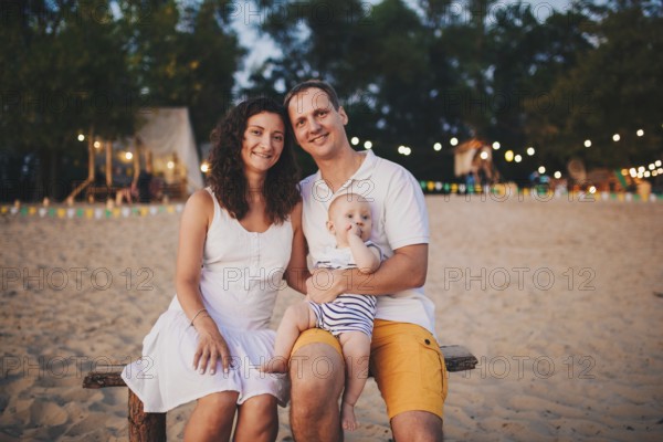 Theme of family summer holidays on the beach. Caucasian man and woman dad and mom hug a small baby son sitting on his hands. Young family relaxing in a beach cafe at sunset