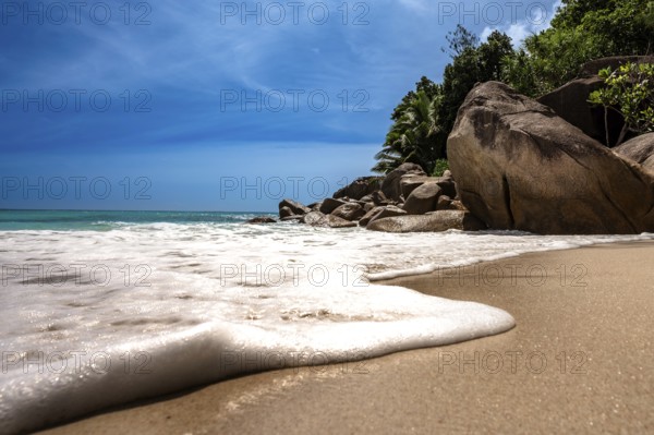 Waves crash over a peaceful tropical beach lined with rocks and trees, La Digue, Seychelles