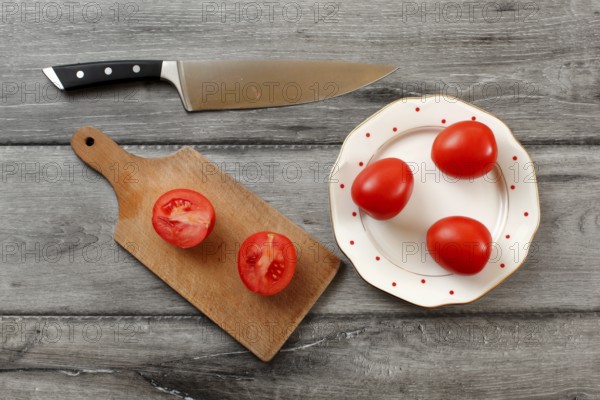 Table top view on three tomatoes on white porcelain plate with red dots, cutting board with tomato cut to half next to it, chef knife also laying on the gray wood desk