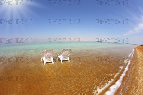 Two white beach chairs stood side by side in the clear water. The sun shines on the beach therapeutic