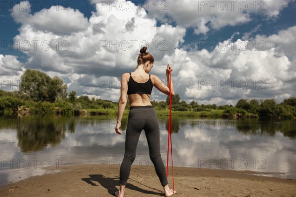 A woman exercises with a resistance band near a calm body of water under a cloud-filled sky