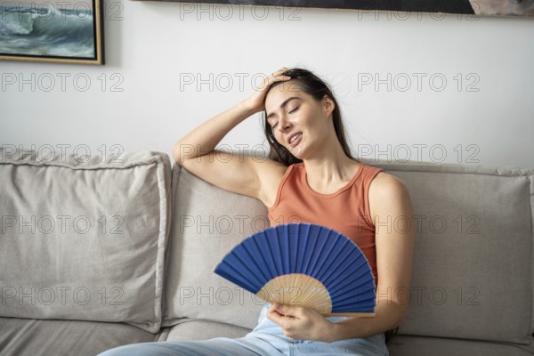 Satisfied woman smiling, blowing with fan, sitting on sofa in home studio. Happy pleased girl feeling comfortable, relieved, cooling down coping with summer heat and stuffiness by fanning