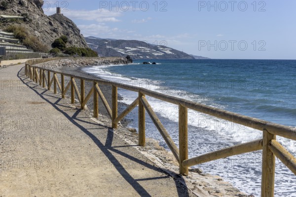 Promenade with wooden railings by the sea in front of the village with blue sky and sea, Castillo de Baños de Abajo, Polopos, Granada, Andalusia, Spain