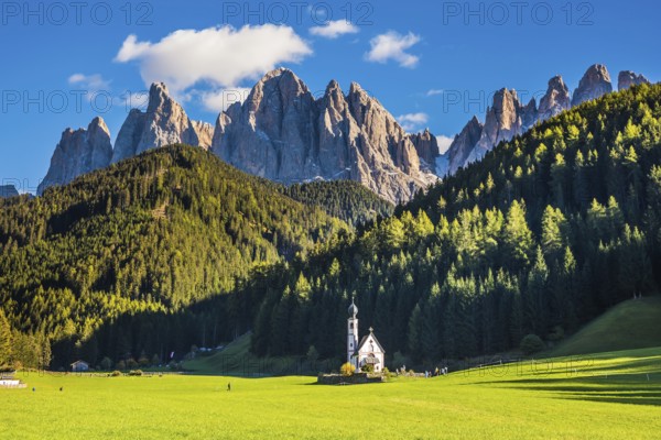 The famous symbol of the valley Val di Funes - church of Santa Maddalena. Rocky peaks and forested mountains surrounded by green Alpine meadows. Dolomites, Tirol. Sunny warm autumn day